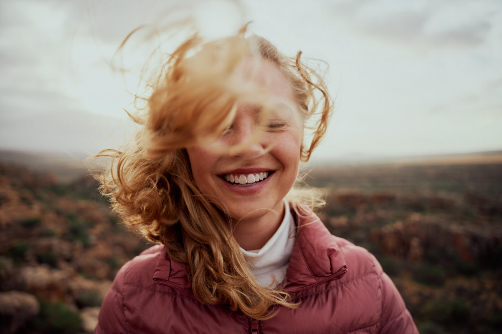 Portrait of young smiling woman face partially covered with flying hair in windy day standing at mountain carefree woman